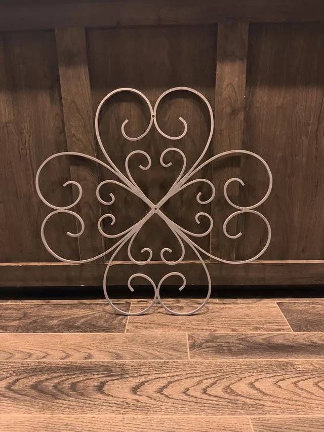 A white scrolled steel wall piece in a four-leaf pattern leaning against a timber wall