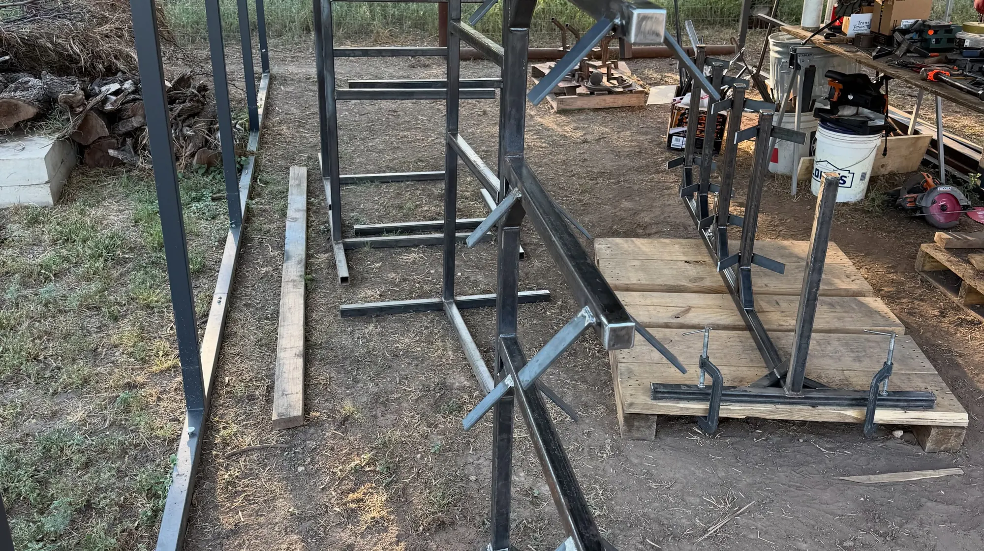 Six steel saddle racks mid-build on pallets and sawhorses in the yard, clamped square, with the welding bench and tools behind them