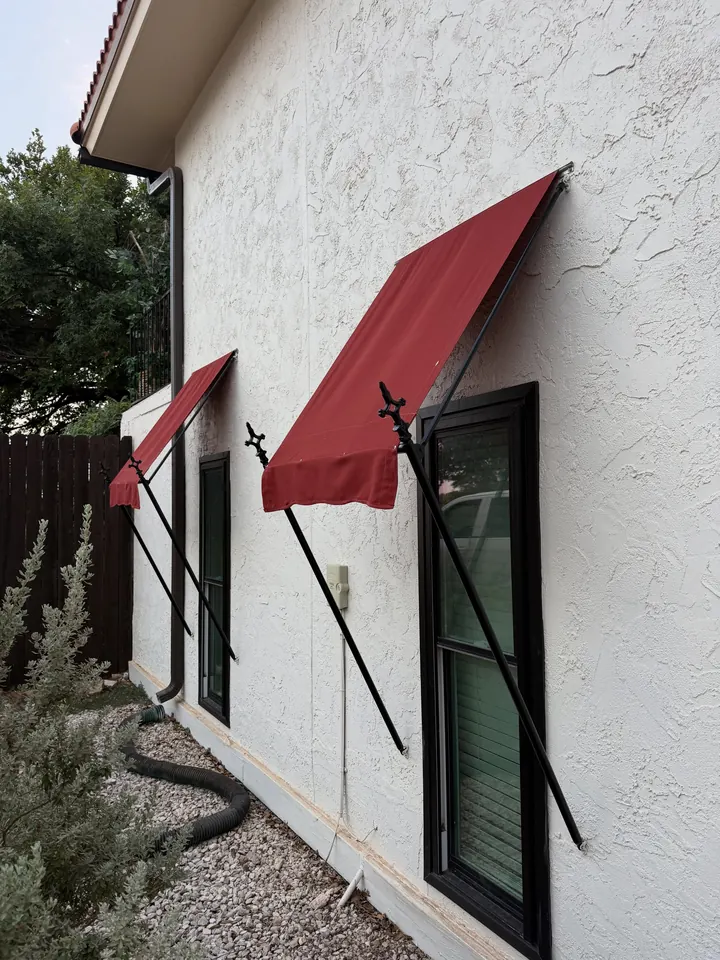 Two red fabric window awnings on steel frames and rods against a white stucco wall