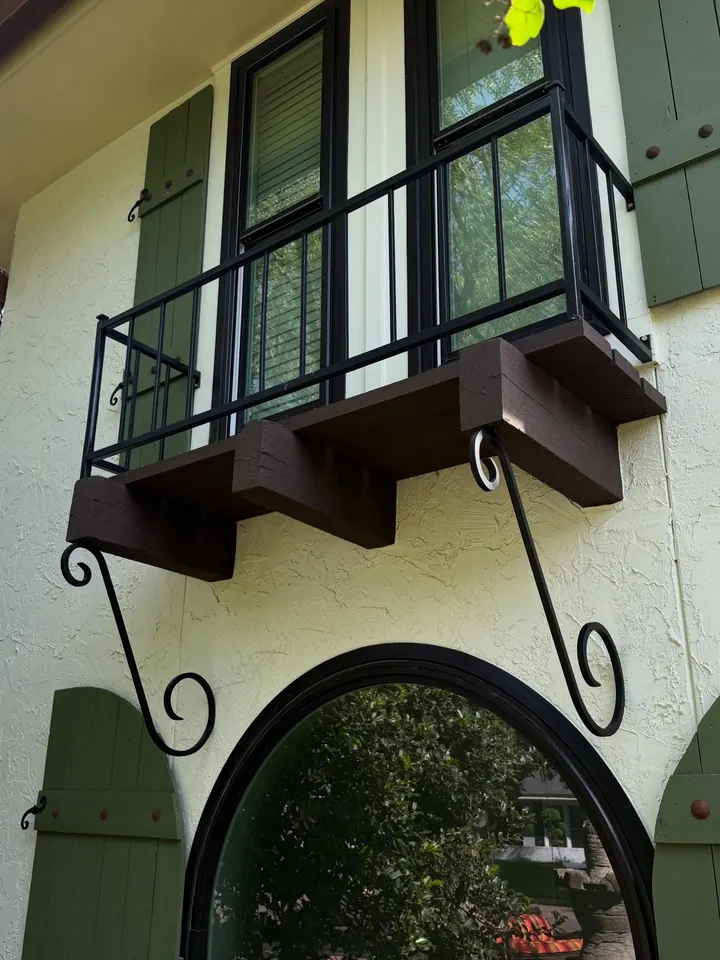 Black steel balcony railing on white stucco, carried on timber joists with scrolled iron brackets curling down the wall