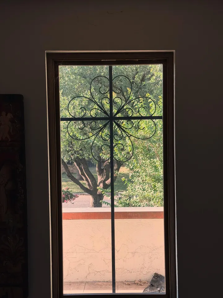 A tall window with a black scrolled iron cross fitted inside the frame, trees beyond the glass